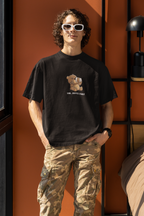 Man wearing a black oversized t-shirt with a cartoon bear drinking coffee and the text “Shh... Coffee First”, wearing white sunglasses ,black watch and standing by an orange wall.