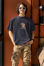 Man wearing a navy blue oversized t-shirt with a cartoon bear drinking coffee and the text “Shh... Coffee First”, wearing white sunglasses ,black watch and standing by an orange wall.