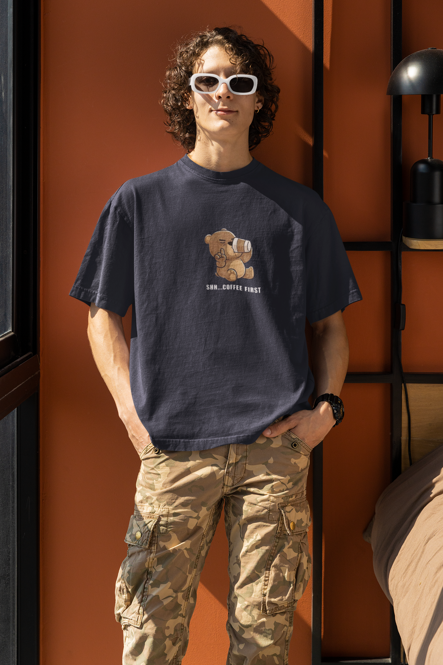 Man wearing a navy blue oversized t-shirt with a cartoon bear drinking coffee and the text “Shh... Coffee First”, wearing white sunglasses ,black watch and standing by an orange wall.