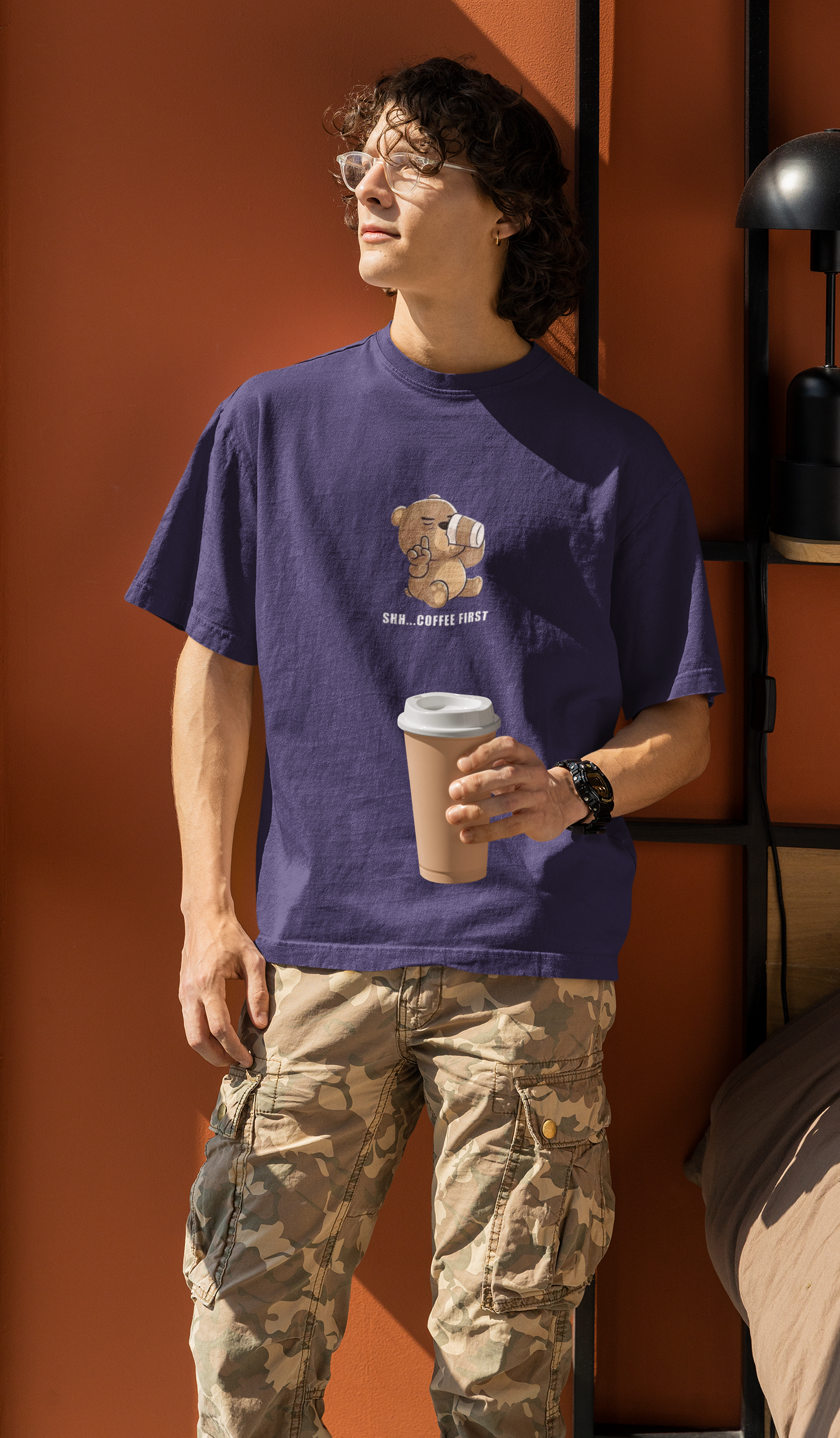 Man wearing a purple oversized t-shirt with a cartoon bear drinking coffee and the text “Shh... Coffee First”, holding a coffee cup and standing by an orange wall.