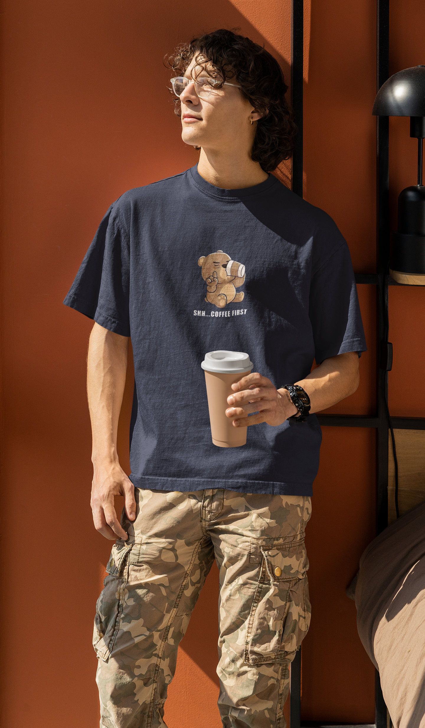 Man wearing a navy blue oversized t-shirt with a cartoon bear drinking coffee and the text “Shh... Coffee First”, holding a coffee cup and standing by an orange wall.