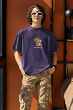 Man wearing a purple oversized t-shirt with a cartoon bear drinking coffee and the text “Shh... Coffee First”, wearing white sunglasses ,black watch and standing by an orange wall.