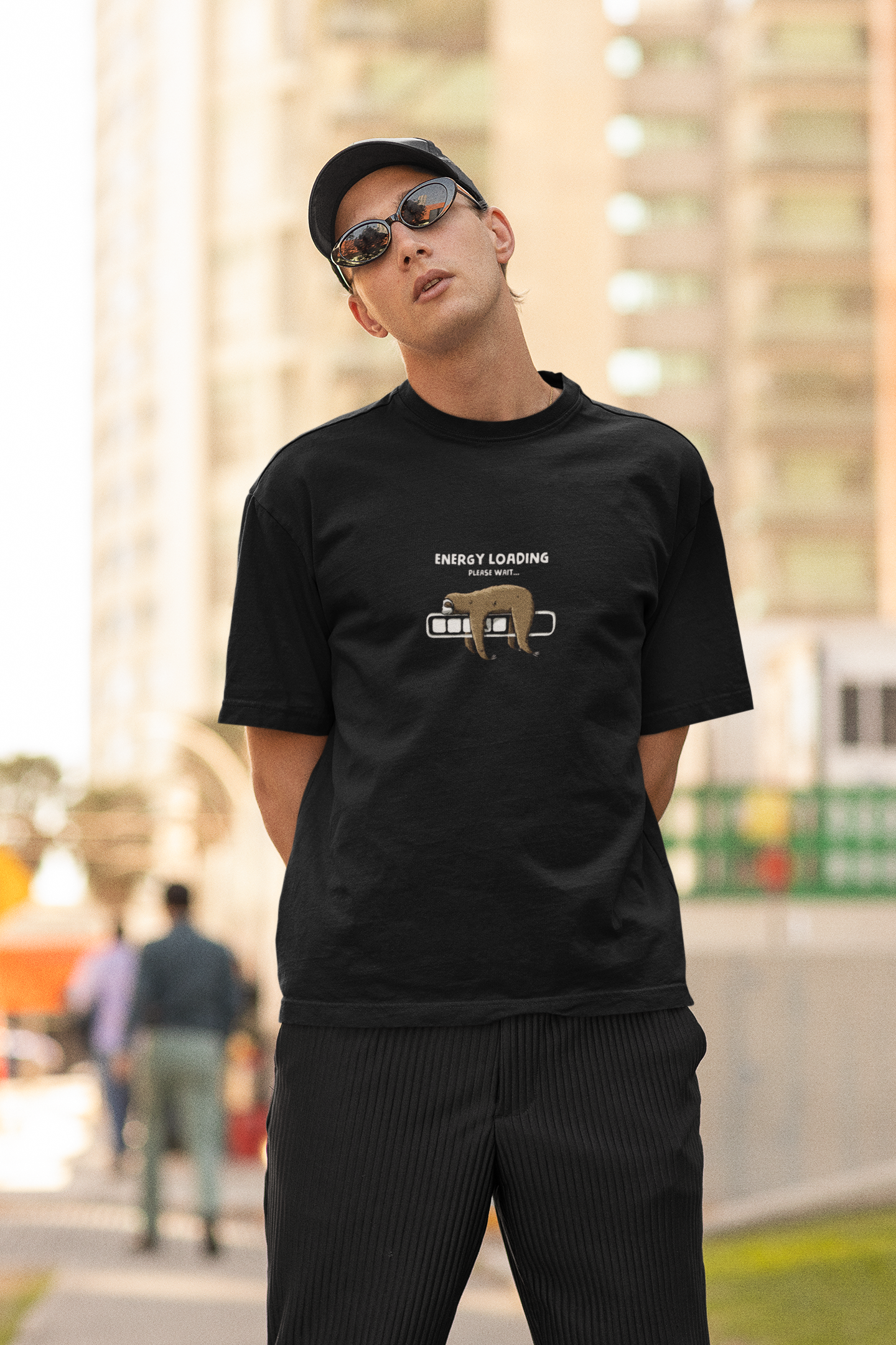 Man wearing a black oversized t-shirt with a sloth and “Energy Loading, Please Wait…” graphic, styled with black cap and sunglasses in an urban outdoor setting.