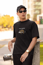 Man wearing a black oversized t-shirt with a sloth and “Energy Loading, Please Wait…” graphic, styled with black cap, sunglasses, and gold watch, in an urban outdoor setting.