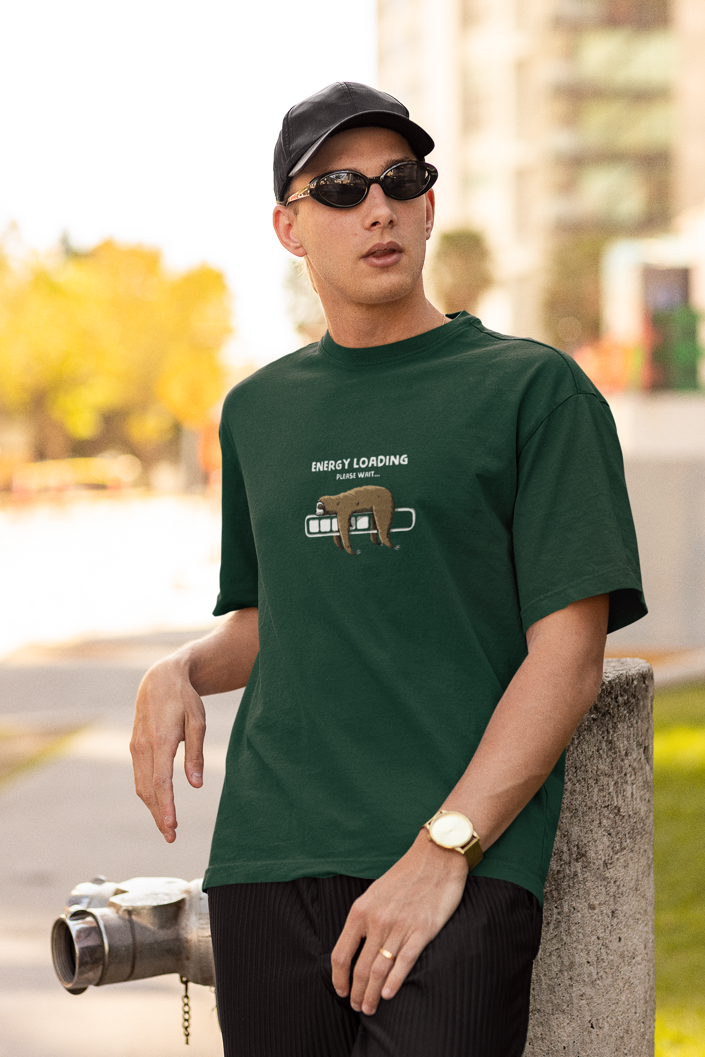 Man wearing a forest green oversized t-shirt with a sloth and “Energy Loading, Please Wait…” graphic, styled with black cap, sunglasses, and gold watch, in an urban outdoor setting.