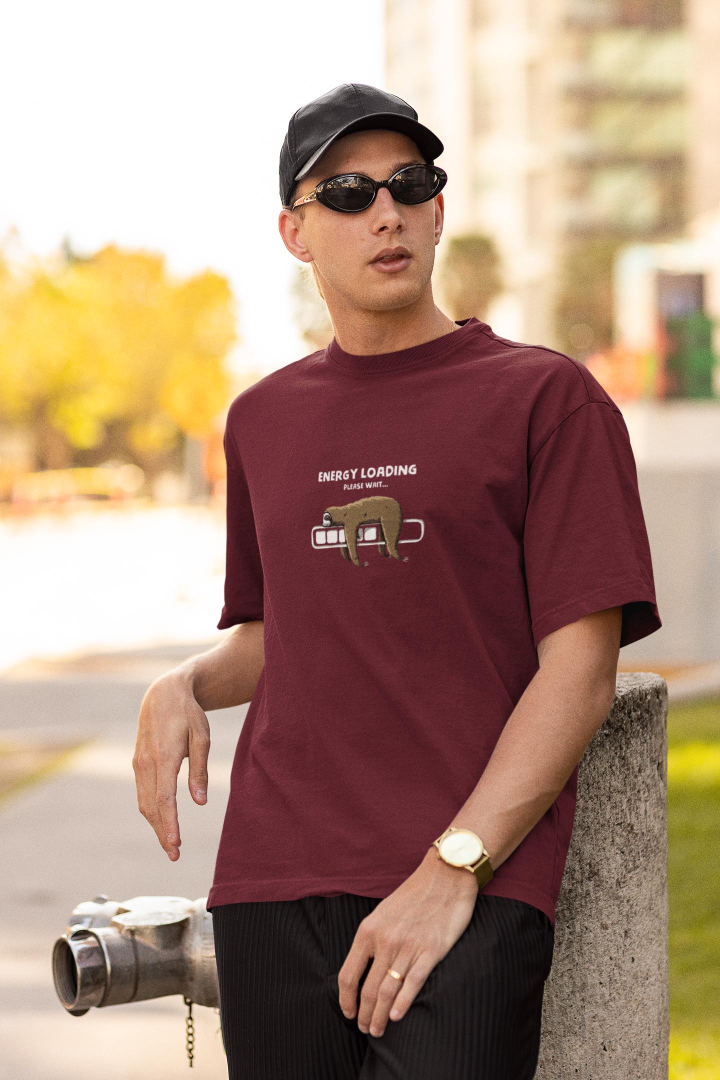 Man wearing a maroon oversized t-shirt with a sloth and “Energy Loading, Please Wait…” graphic, styled with black cap, sunglasses, and gold watch, in an urban outdoor setting.