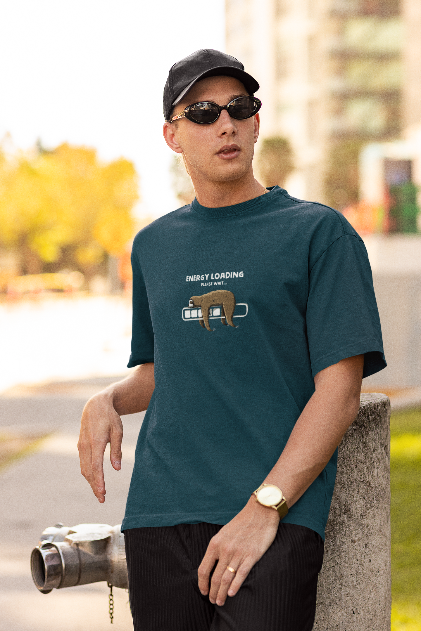 Man wearing a petrol blue oversized t-shirt with a sloth and “Energy Loading, Please Wait…” graphic, styled with black cap, sunglasses, and gold watch, in an urban outdoor setting.