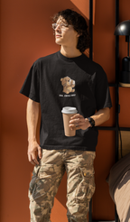 Man wearing a black oversized t-shirt with a cartoon bear drinking coffee and the text “Shh... Coffee First”, holding a coffee cup and standing by an orange wall.